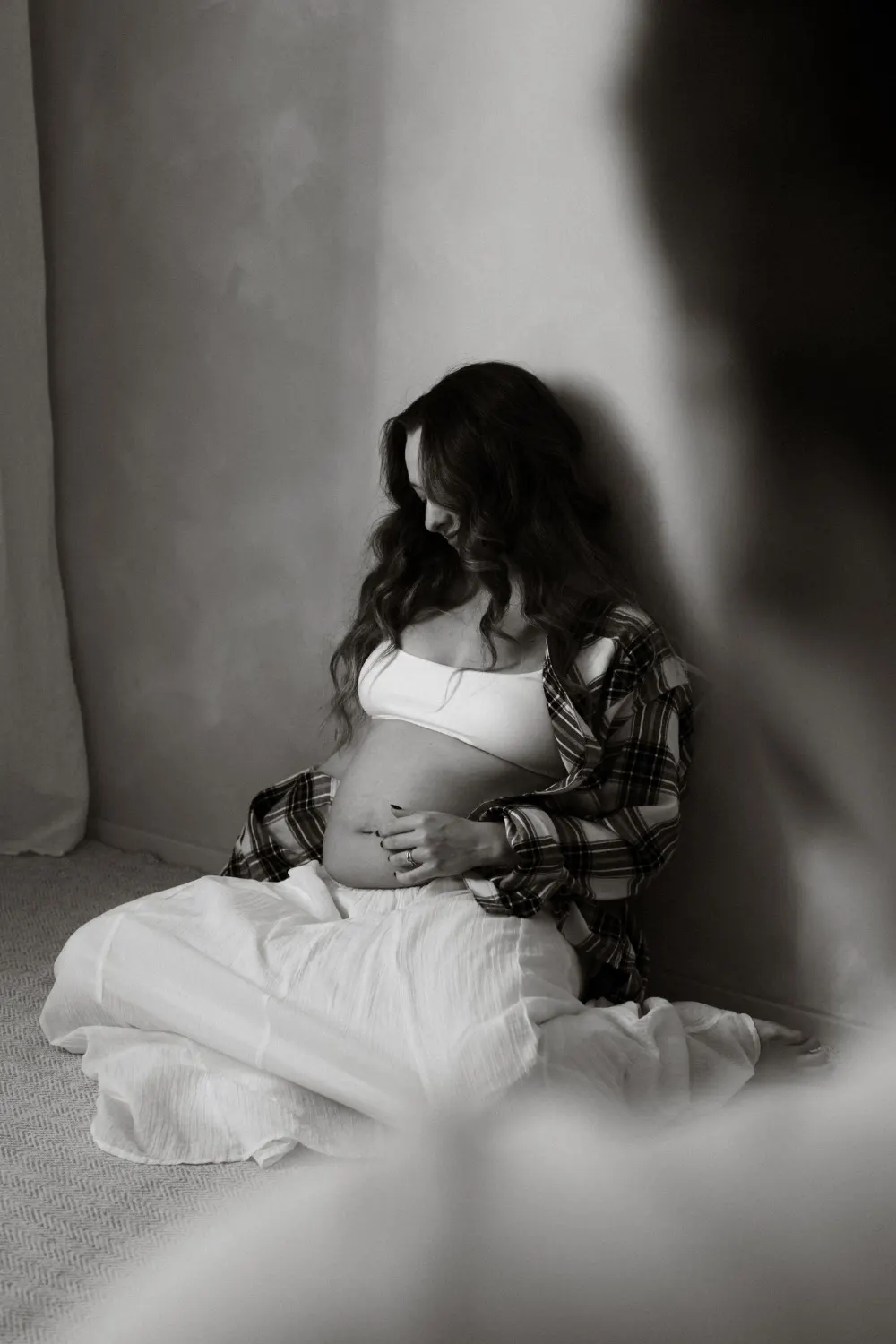 Těhotná žena sedí na zemi v kostkované košili a sukni s odhaleným bříškem při focení v ateliéru