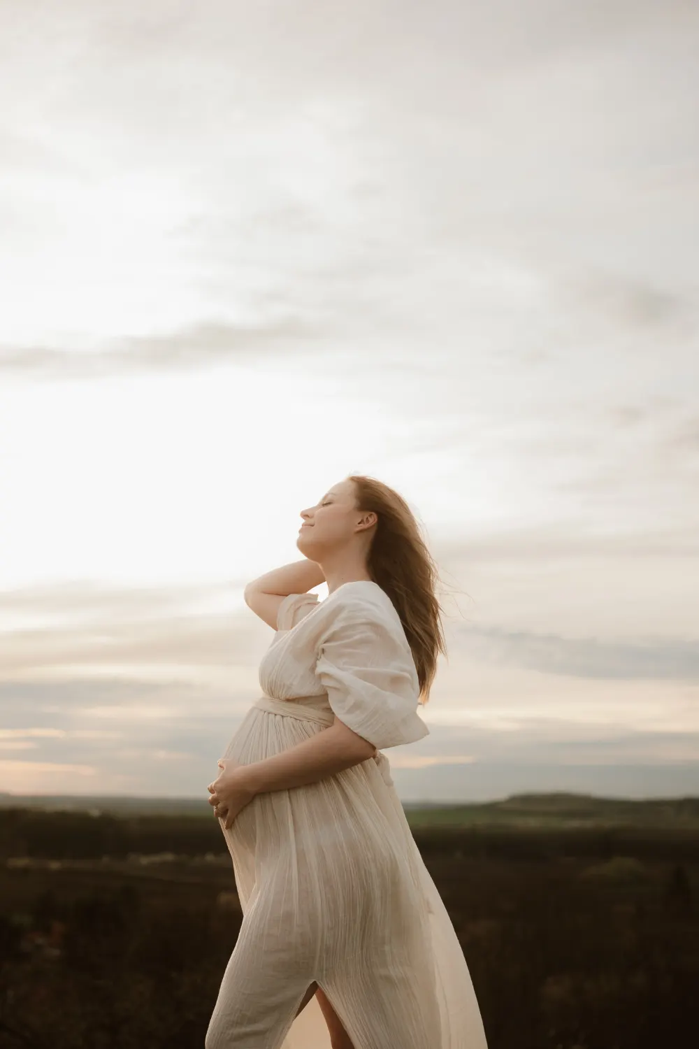 těhotná žena v bílých boho šatech stojí na vrcholu kopce a prohrabává si vlasy