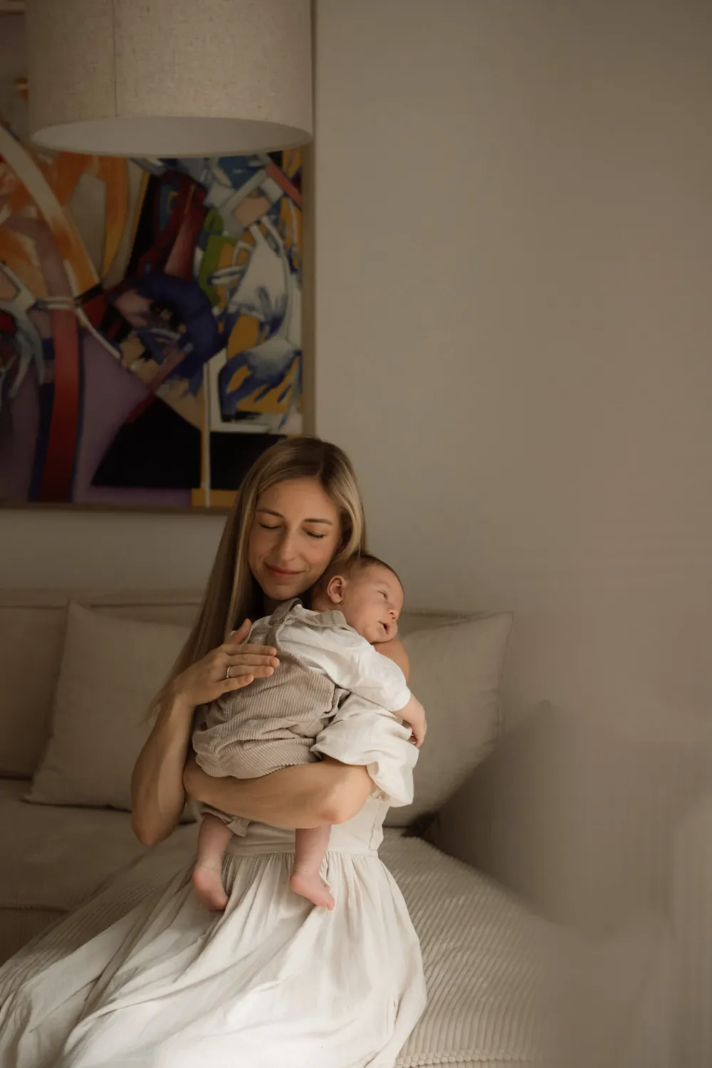 maminka sedí na béžové sedačce a chová svého novorozence na rameni