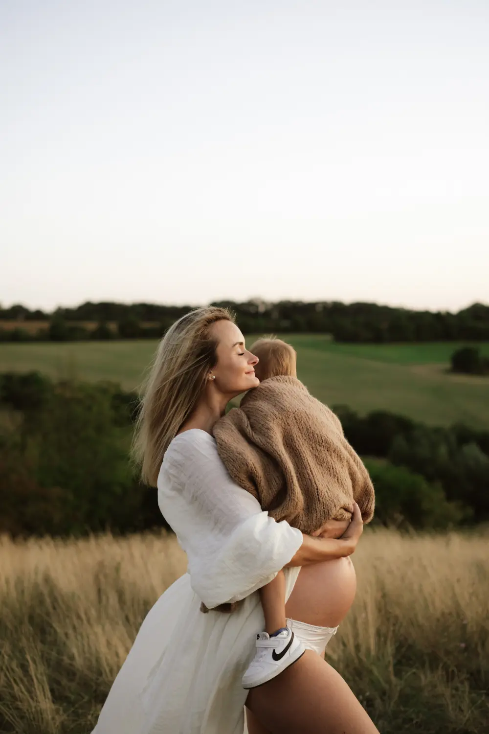 těhotná žena v bílých šatech drží v náručí syna a objímají se na kopci